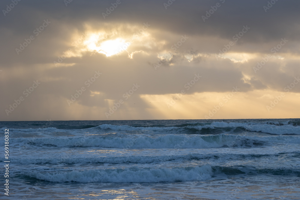 Obraz premium Foamy sea waves and sun peaking through the clouds at early evening