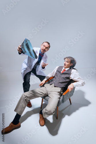 Emotional doctor showing X-ray examination to scared young man sitting on chair. Medical check up. Models against grey studio background. Concept of profession, occupation, medicine, emotions