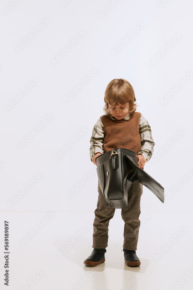 Serious little boy, kid wearing retro style clothes looking into retro suitcase isolated over white background. Concept of emotions, 50s, 60s vintage fashion style, childhood Serious little boy, kid wearing retro style clothes looking into retro suitcase isolated over white background. Concept of emotions, 50s, 60s vintage fashion style, childhood