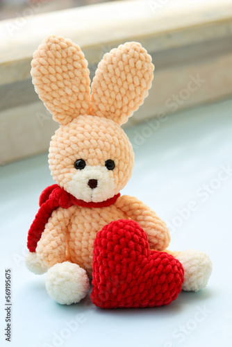 Saint Valentine's day creative image with a handmade teddy rabbit sitting with a big knitted red heart as a present. Self made knitted rabbit toy in pastel colors holding a red heart. Love and couple