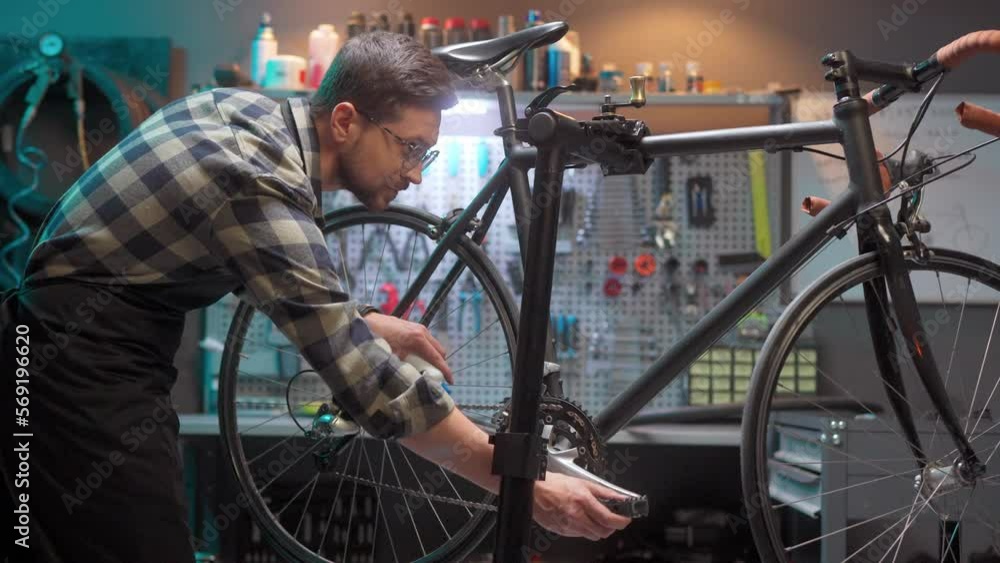 Handsome caring skillful man repairing and maintaining bicycle in modern authentic garage lubricating bike chain. Beautiful focused male repairman doing hobby fixing bikes wearing glasses. Handsome caring skillful man repairing and maintaining bicycle in modern authentic garage lubricating bike chain. Beautiful focused male repairman doing hobby fixing bikes wearing glasses.