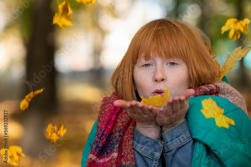 Red-haired girl in the autumn park blowing on maple leaves.