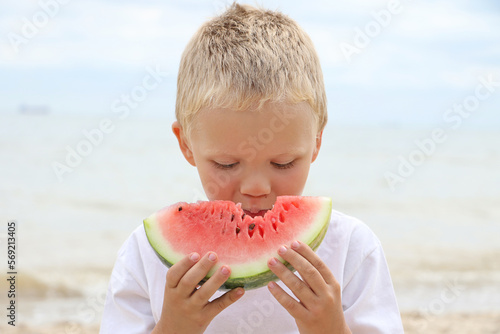 Sweet sun tanned blonde kid on the coast, holding juicy slice of the watermelon and eat it.Summertime concept.