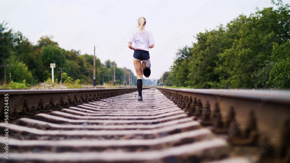 Beautiful sexy athletic young blond woman in top and shorts, running, jogging, Rear view. On the railway, on rails, sleepers, in the summer.