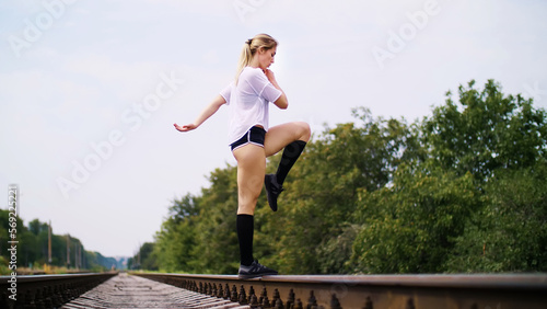 Beautiful sexy athletic young blond woman in top and shorts, performs exercises for legs, lifting legs with a bent knee, On railway, on rails, sleepers, in summer. slow motion