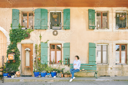 Young pregnant woman sitting on bench next to beautiful village house facade