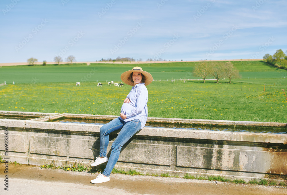 Outdoor portrait of happy young pregnant woman enjoying nice day in countryside, sitting on watering fountain, green pasture with cows on background