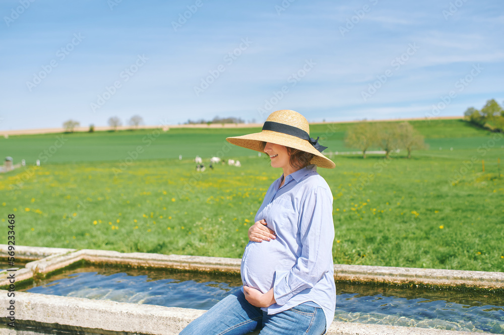 Outdoor portrait of happy young pregnant woman enjoying nice day in countryside, sitting on watering fountain, green pasture with cows on background