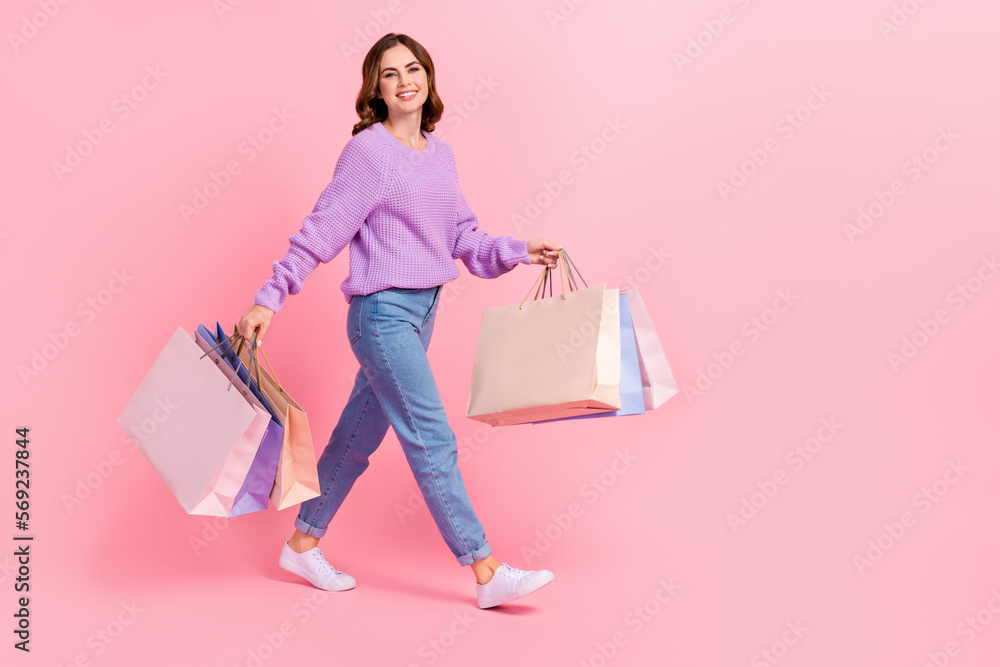 Full length photo of cute shiny girl dressed purple pullover walking holding shoppers empty space isolated pink color background
