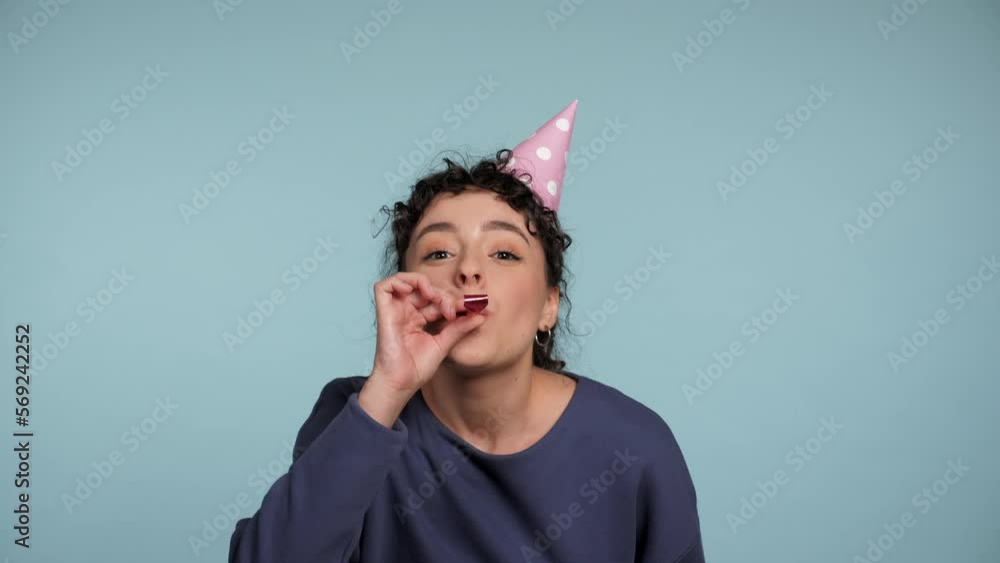 Close up happy curly woman in birthday cap blowing whistle celebrating holiday on isolated light blue background with copy space. Positive smiling cute female wearing blue sweater. People emotions Close up happy curly woman in birthday cap blowing whistle celebrating holiday on isolated light blue background with copy space. Positive smiling cute female wearing blue sweater. People emotions
