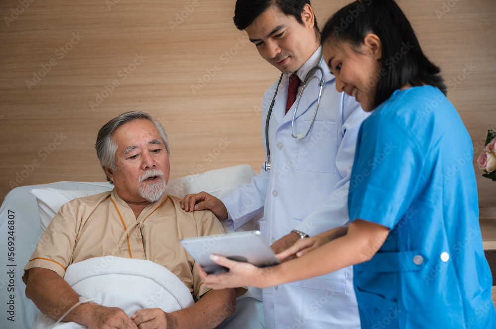 Fototapeta premium Smiling senior male patient lying and sitting on bed resting while nurse and doctor in lab coat and stethoscope showing digital tablet screen to man in a hospital room