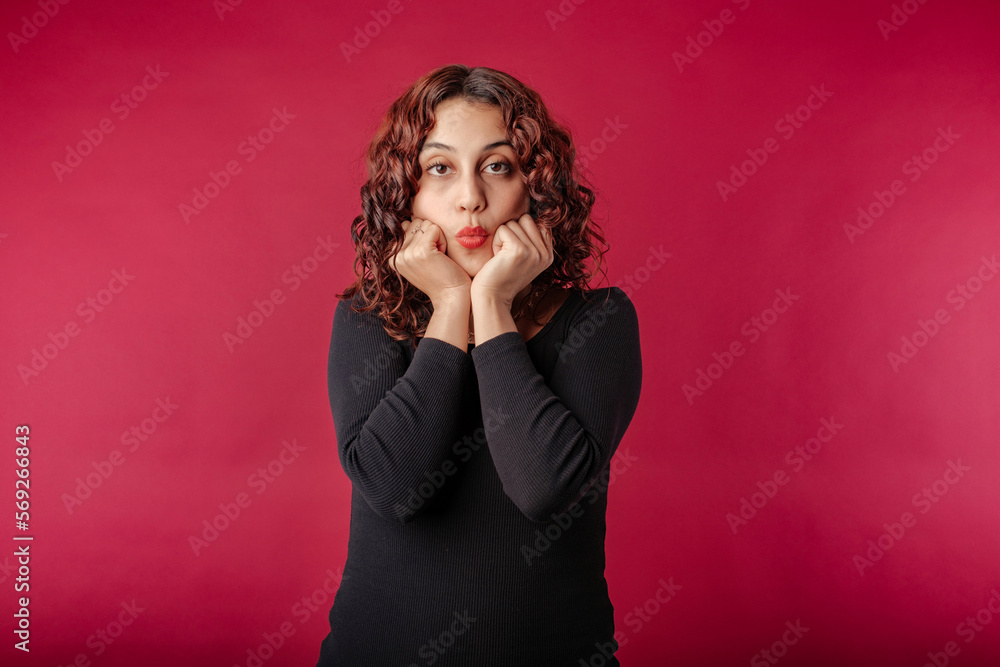 Portrait of cheerful woman wearing dress standing isolated over red background hands in fists, touching chin, looks wow mouth at the camera and is dumbfounded. Can't wait to share what has heard. Portrait of cheerful woman wearing dress standing isolated over red background hands in fists, touching chin, looks wow mouth at the camera and is dumbfounded. Can't wait to share what has heard.