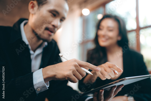 Smiling and laughing young Asian successful business woman discussing work with male colleague and partner in modern office while using digital tablet