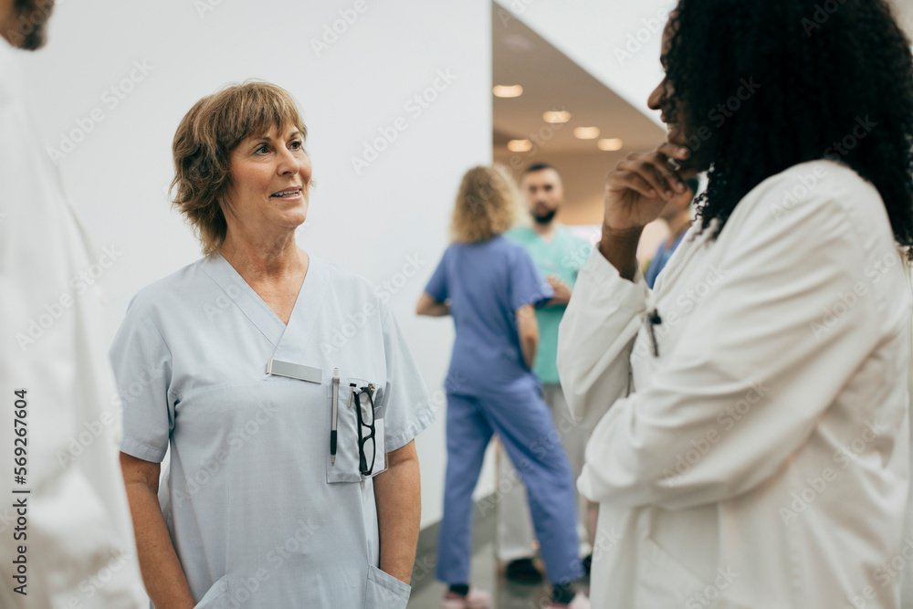 Smiling female doctor with hands in pockets discussing with colleague at hospital Smiling female doctor with hands in pockets discussing with colleague at hospital