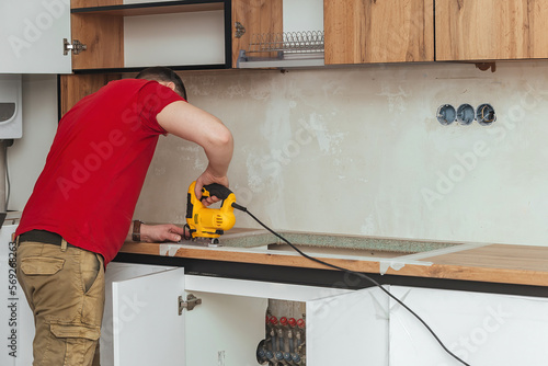 The furniture master installs a modern kitchen in the house, in his hands is an electric jigsaw with which he cuts a place for a sink in the countertop.