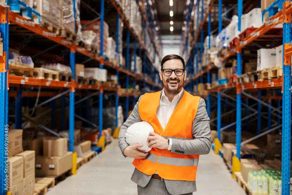 A businessman is standing with helmet under armpit in storage and smiling at the camera.