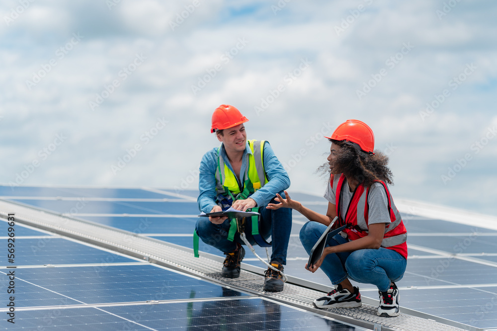 Two young engineer and inspector wearing safety vest and hard helmet inspecting solar panel by kneeling and filling holes and fixing damage using a drill machine while holding a clipboard Two young engineer and inspector wearing safety vest and hard helmet inspecting solar panel by kneeling and filling holes and fixing damage using a drill machine while holding a clipboard