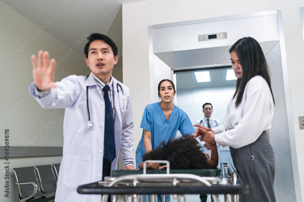 Fototapeta premium Portrait of young handsome Asian male doctor rushing in hospital during emergency with patient lying on bed with mother holding hand and nurse assisting in corridor passage