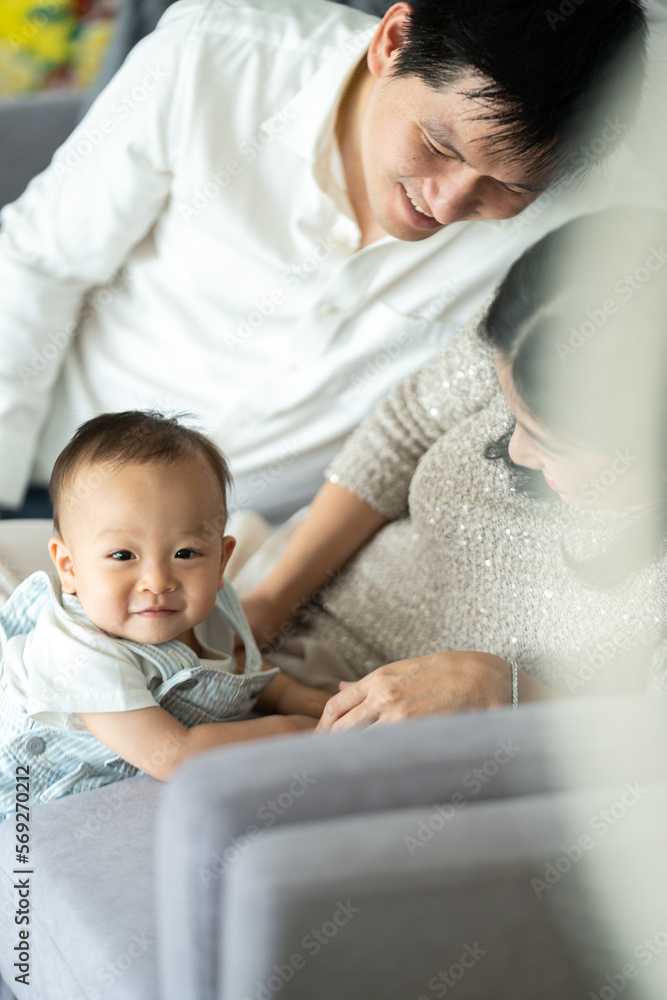 Young and cheerful mother and father sitting on couch relaxing while playing with cute little new born baby boy and toddler who is trying to stand with support at home during weekend Young and cheerful mother and father sitting on couch relaxing while playing with cute little new born baby boy and toddler who is trying to stand with support at home during weekend