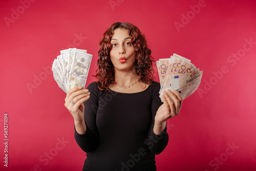 Redhead millennial woman wearing black dress standing isolated over red background holding a wad of cash looks at the camera, scared and amazed and open mouth. Unbelievable. So much money.