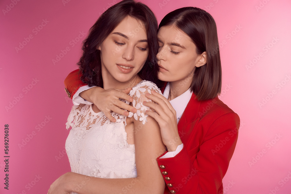 Valentine's day concept. Studio portrait of two young lesbian girls. One girl is in a white dress, the second woman is in a red suit. They stand on a pink background. Cropped shot. Valentine's day concept. Studio portrait of two young lesbian girls. One girl is in a white dress, the second woman is in a red suit. They stand on a pink background. Cropped shot.