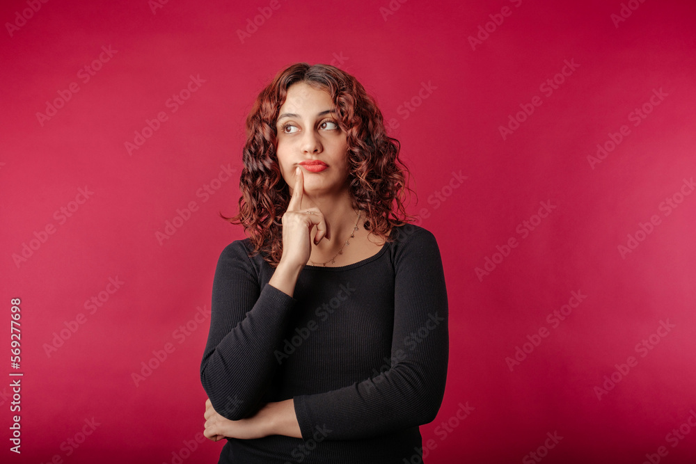 Fototapeta premium Young beautiful woman wearing black ribbed dress isolated over red background undecided, purses lips and touches with index finger. Looks at the empty copy space and tries to decide.