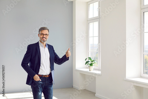 Joyful male realtor or real estate agent showing thumb up while standing in new building. Man in suit stands in bright room and recommends his services. Real estate business, sale and people concept.