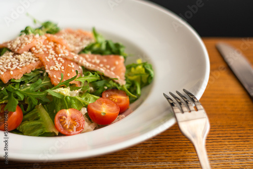 White ceramic plate of fresh salad with salmon, vegetables, tomatoes and arugula with sesame seeds.