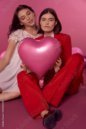 Valentine's day concept. Studio portrait of a lgbtq couple holding a pink heart balloon and sitting on the floor on a pink background. One girl is in a white dress, the second woman is in a red suit.