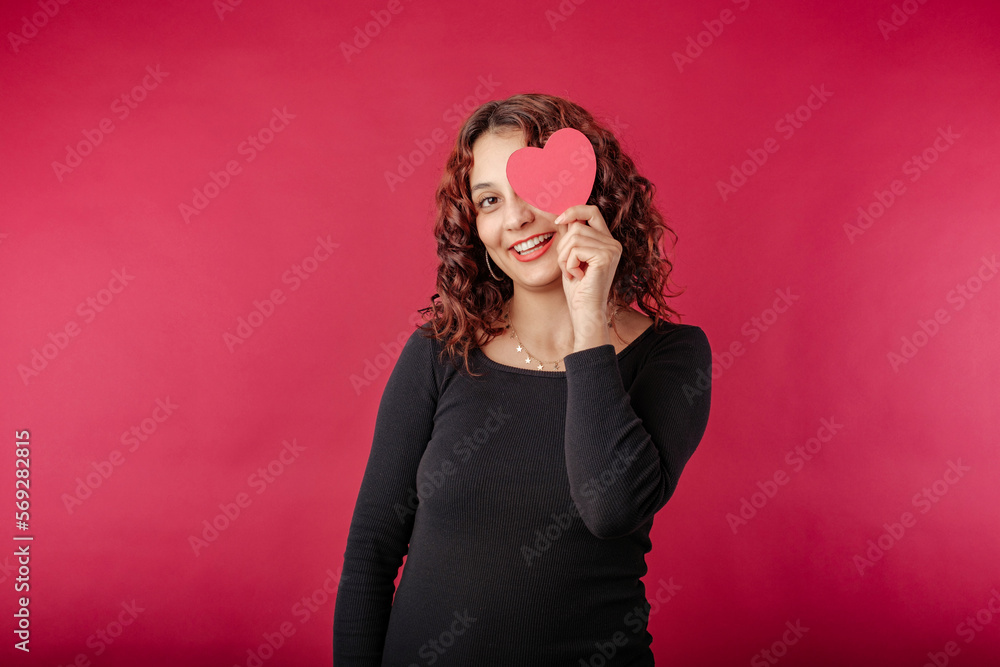 Φωτογραφία Cheerful redhead woman wearing black ribbed dress isolated over red background covering one eye with a small cardboard heart shape, laughs and looks at the camera