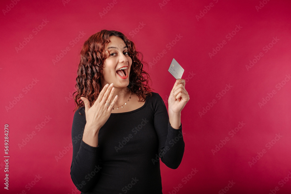 Happy redhead woman wearing black ribbed dress standing isolated over red background holding credit card, showing membership card mockup shopping payment money financial online.