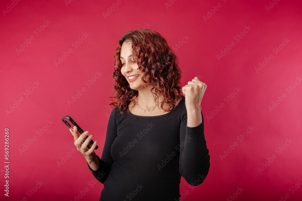 Portrait of cute redhead woman wearing black ribbed dress standing isolated over red background holding the phone and makes a fist while looking at the phone and makes a gesture of joy or winning. Portrait of cute redhead woman wearing black ribbed dress standing isolated over red background holding the phone and makes a fist while looking at the phone and makes a gesture of joy or winning.