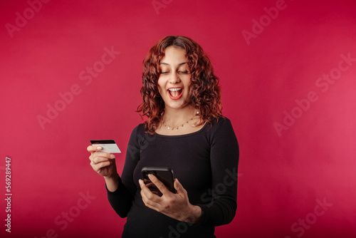 Happy redhead woman standing isolated over red background holding a credit card and makes a gesture of joy while looking at the phone screen. Cannot believe what sees on the screen.