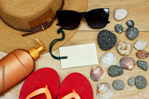 orange beach slippers and hat, sunglasses, bottle of cream sun with seashells and stones, empty tag on wooden table