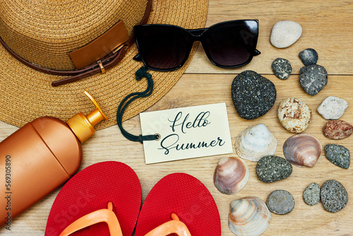 orange beach slippers and hat, sunglasses, bottle of cream sun with seashells and stones, hello summer text tag on wooden table