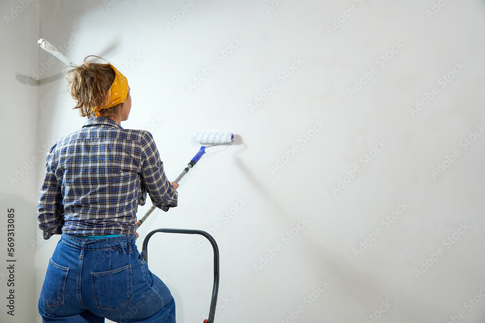 Positive overjoyed woman painter holding roller painter painting walls in room. People renovation and home improvement concept. View from the back Positive overjoyed woman painter holding roller painter painting walls in room. People renovation and home improvement concept. View from the back