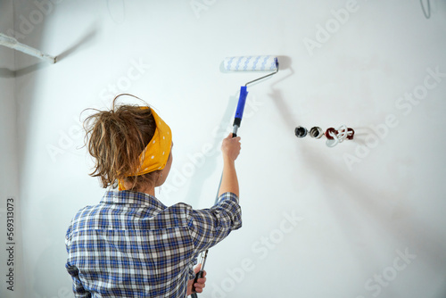 Positive overjoyed woman painter holding roller painter painting walls in room. People renovation and home improvement concept. View from the back