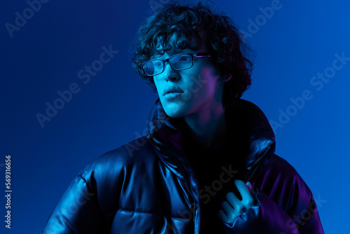 Man in winter jacket model poses in glasses smile, hipster lifestyle, portrait blue background, mixed neon light, fashion style and trends boys teenager, copy space