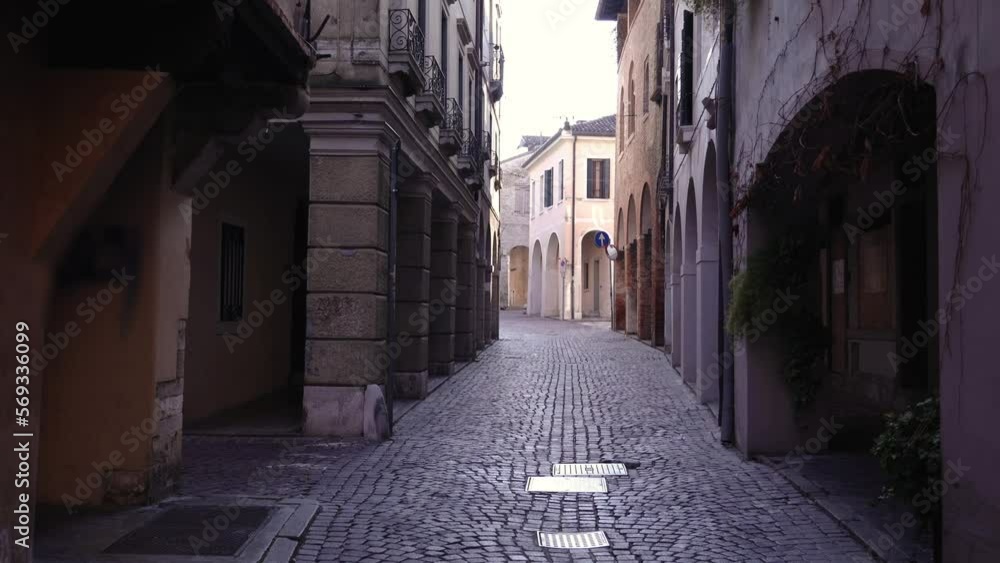 Historical city buildings with large brick arches and grey paved road on empty narrow shadow street leading to sunny square