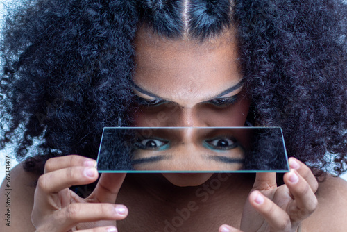Mujer joven latina con el pelo rizado y un vestido de tirantes de varios colores posando delante de un espejo con el fondo una pared blanca.