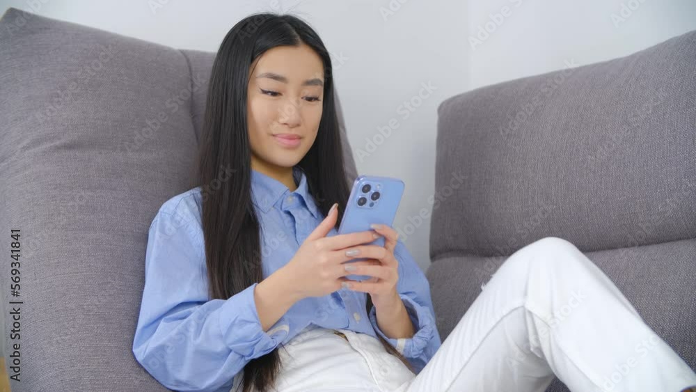 Beautiful Vietnamese POC female sitting on couch at home and browsing a mobile app on a smartphone. Cute young Asian BIPOC girl using modern mobile phone application with a smile Beautiful Vietnamese POC female sitting on couch at home and browsing a mobile app on a smartphone. Cute young Asian BIPOC girl using modern mobile phone application with a smile
