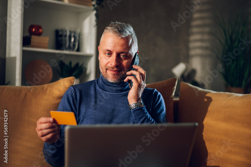 Middle aged man using a smartphone and a laptop while sitting on a sofa at home