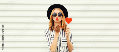 Portrait of beautiful young woman with red heart shaped lollipop blowing her lips with lipstick sending sweet air kiss wearing sunglasses, black round hat on white background
