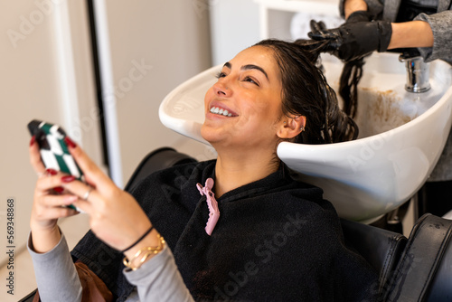 High angle of positive young female customer with smartphone smiling and lying in sink while getting professional procedure at hairdressing salon
