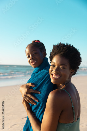 Portrait of smiling african american mother covering daughter with towel at beach against clear sky