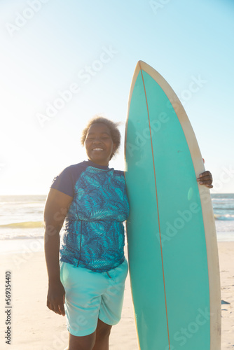 African american senior woman with surfboard standing at beach against sea and sky at sunset