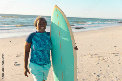 Cheerful african american senior woman with surfboard standing at beach against sea and clear sky