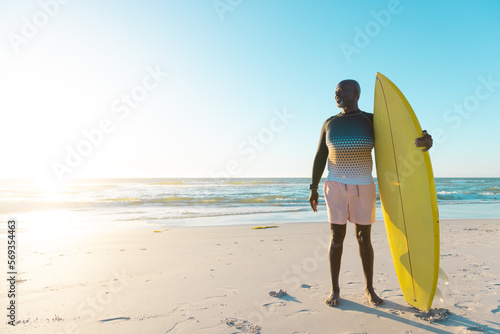 African american senior man with yellow surfboard standing on sandy beach against sea and clear sky
