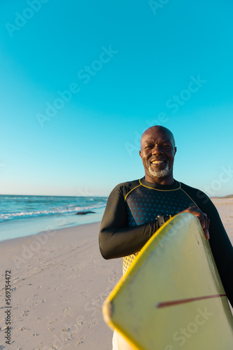 Smiling bald african american senior man carrying surfboard standing at beach against clear blue sky
