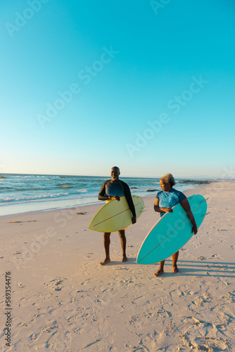 African american senior couple with surfboards walking at sandy beach against sea and blue sky